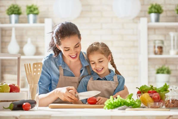 Eine Mutter steht mit ihrer Tochter in der Küche. Beide schneiden gemeinsam eine Tomate und lächeln dabei.