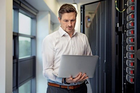 Ein etwa 40 jähriger Mann in weißem Hemd steht mit einem Laptop in der Hand vor einem Server.