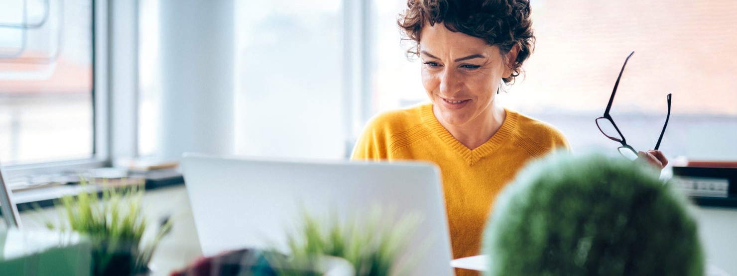 Eine brünette Frau hat eine Brille in der linken Hand, sitzt an einem Schreibtisch und schaut auf Ihren Laptop.