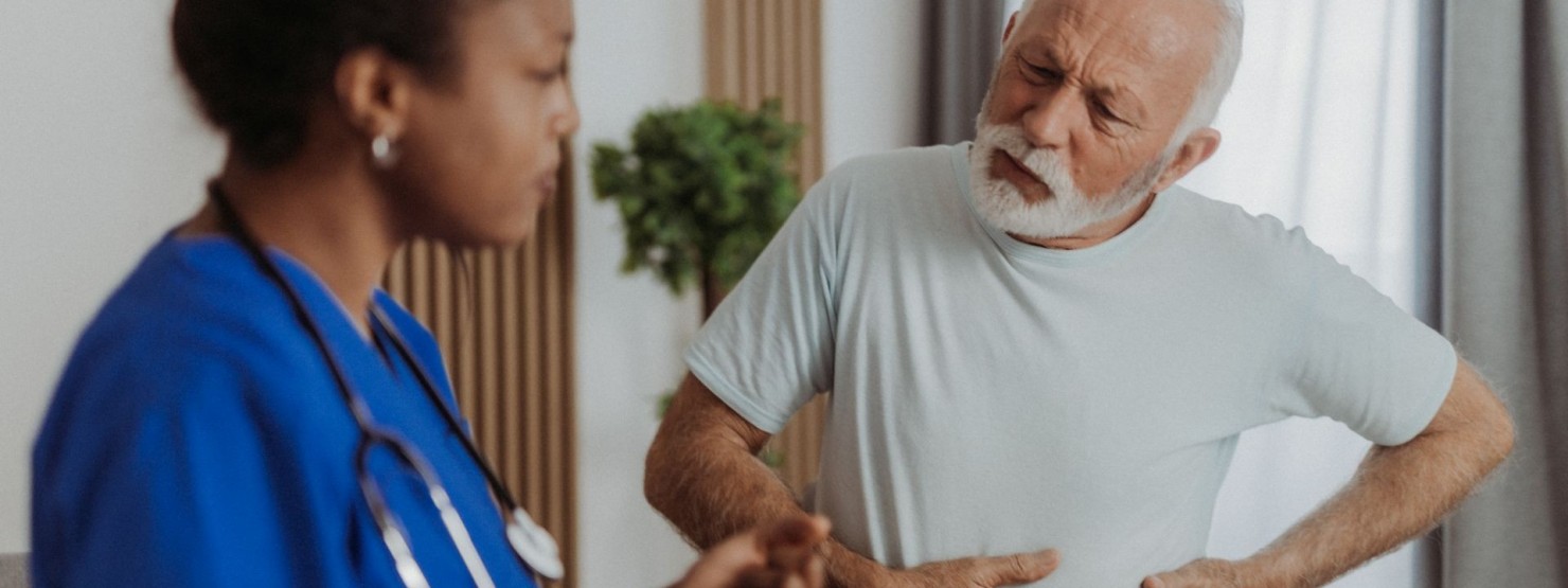 Ein etwa 60-jähriger Mann steht seiner Ärztin. Im Gespräch zeigt er, wo seine Beschwerden im Bauch liegen.