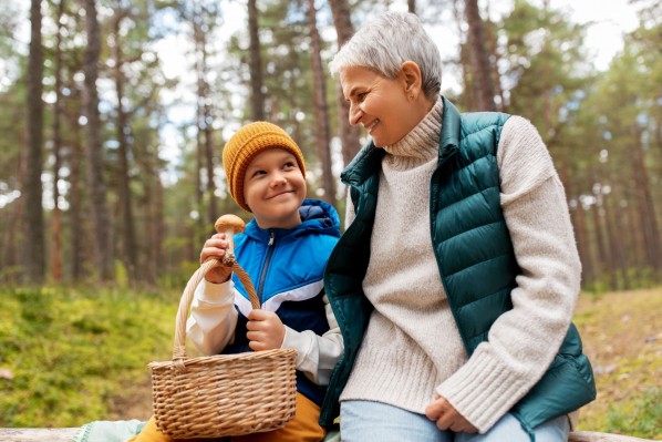 Eine Seniorin sitzt mit ihrem Enkel im Wald. Beide schauen sich an und lächeln. Der Junge zeigt seiner Oma einen Pilz, den er gesammelt hat.