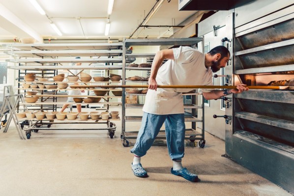 Im Produktionsraum einer kleinen Bäckerei schiebt ein Mann ein Blech voller Brote in den Ofen.