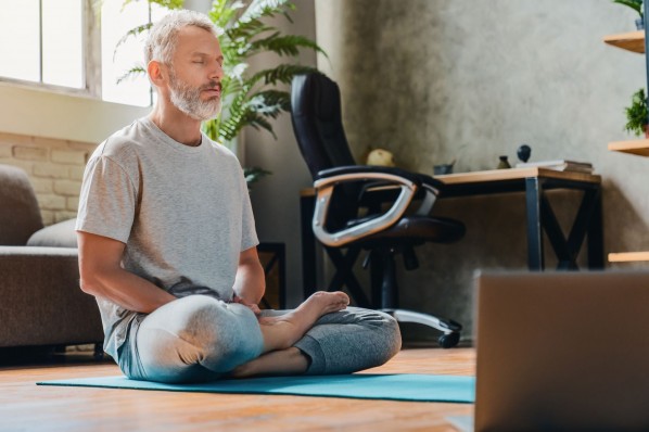 Ein etwa 55 jähriger Mann sitzt im Wohnzimmer im Schneidersitz auf dem Boden auf einer Yogamatte. Er hat die Augen geschlossen und entspannt.