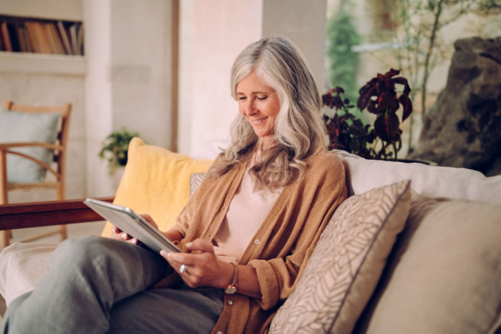 Eine Seniorin sitzt auf ihrem Sofa und schaut glücklich auf ihre Tablet.