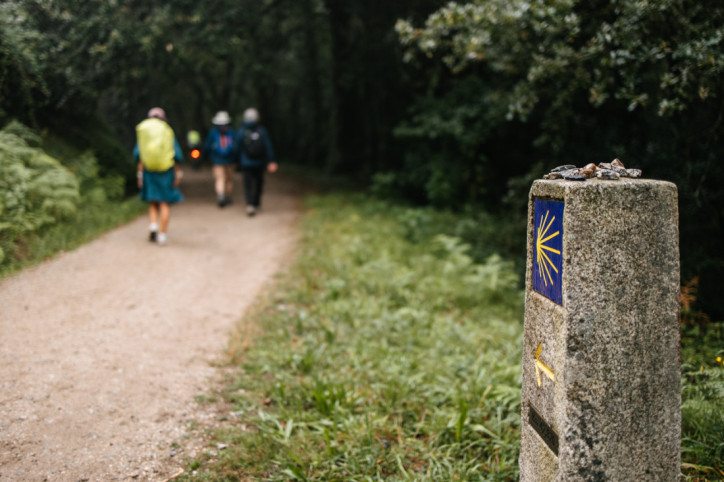 Wegmarkierung Jakobsweg, im Hintergrund drei Pilger mit Rucksack und Hut