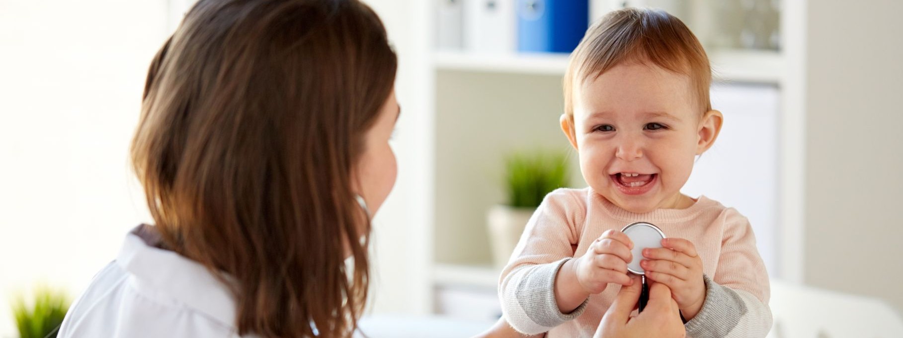 Ein etwa einjähriges Kind wird von einer Kinderärztin mit dem Stethoskop untersucht. Das Kleinkind lacht ausgiebig.