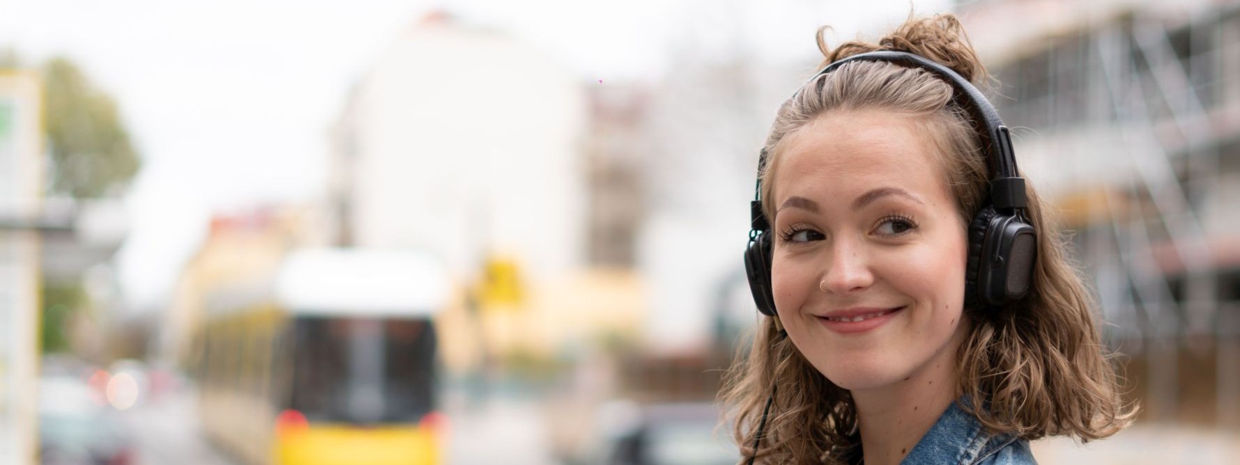 Eine etwa 20 jährige Frau steht an einer Straße und hört über Kopfhörer Musik von ihrem Handy.