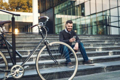 Ein etwas 30 jähriger Mann sitzt auf einer Treppe vor einem Bürogebäude. Er schaut auf sein Handy und lächelt. Vor ihm steht ein Fahrrad.