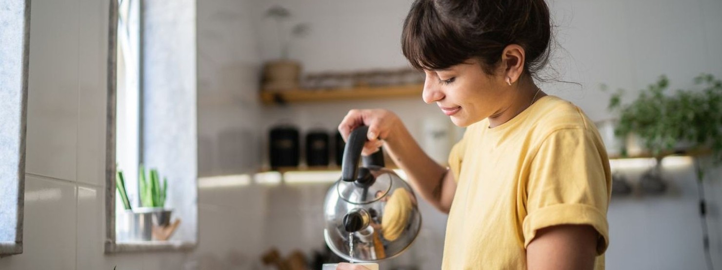 Eine junge Frau steht in ihrer Küche und gießt sich heißes Wasser in eine Tasse. 