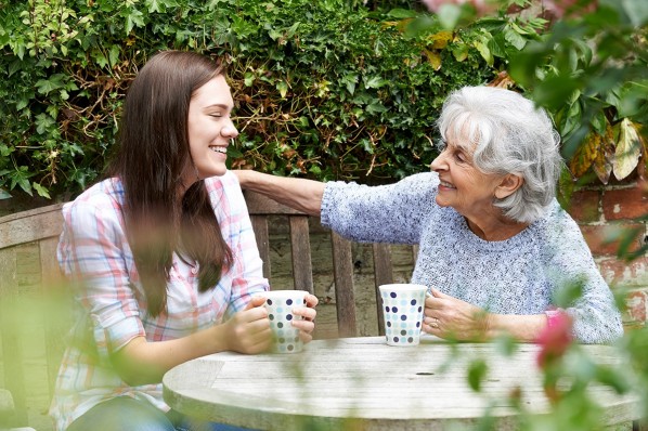 Eine etwa 20-jährige Frau und ihre Oma sitzen mit Tassen im Garten am Tisch.