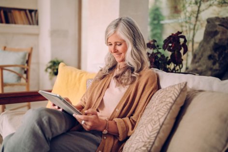 Eine Seniorin sitzt auf ihrem Sofa und schaut glücklich auf ihre Tablet.