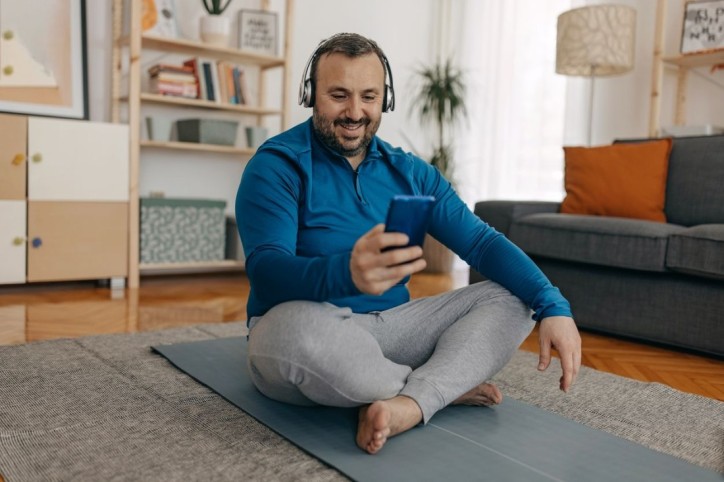 Kräftiger Mann sitzt auf einer Yogamatte und schaut auf sein Handy. Er hat Kopfhörer auf.