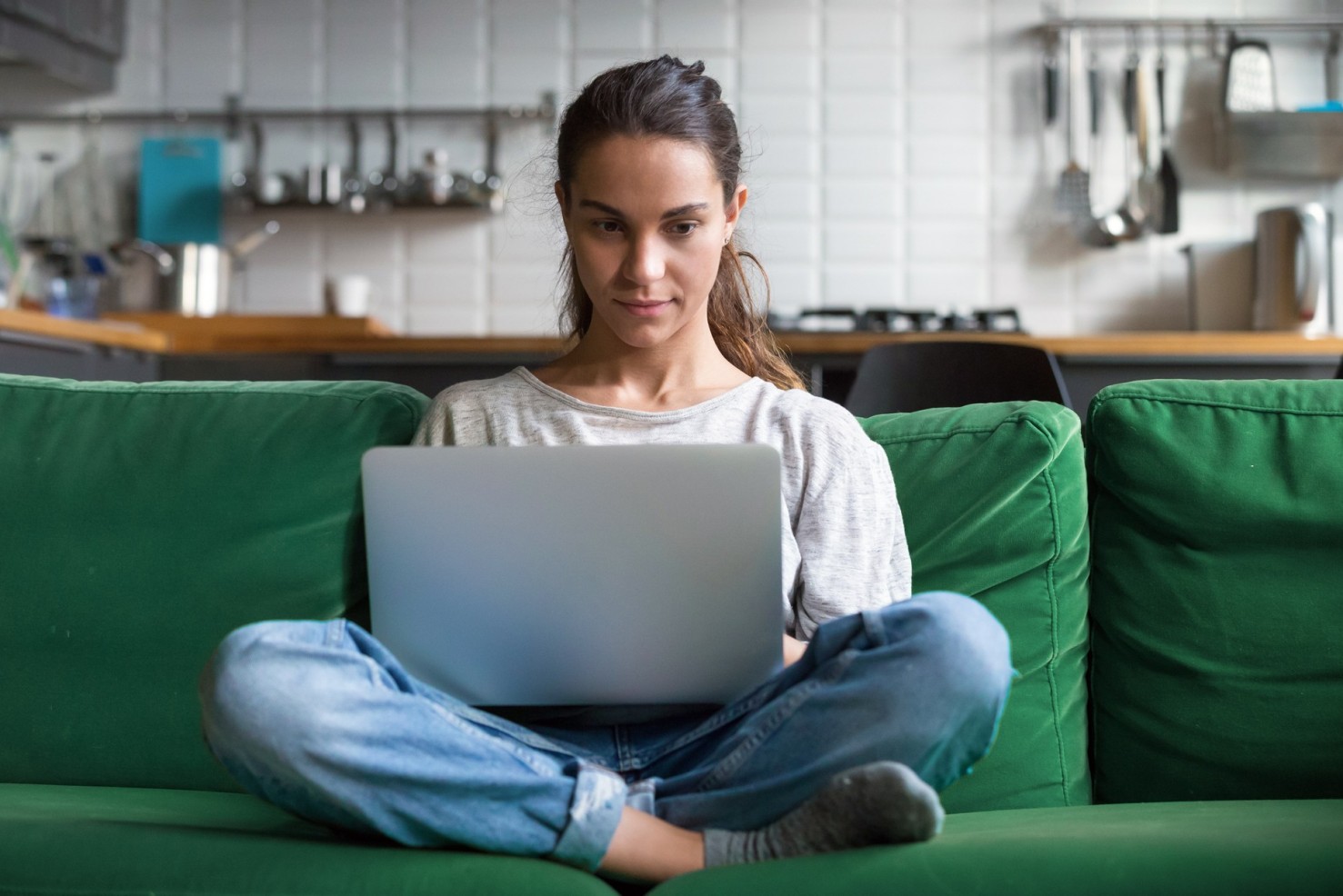 Eine etwa 30-jährige Frau sitz mit einem Laptop auf dem Schoß auf ihrem Sofa