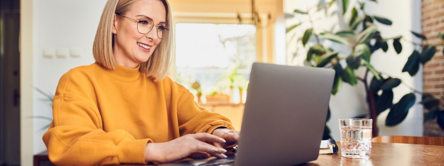 Eine etwa 35-jährige Frau sitzt an einem Tisch und schaut auf Ihren Laptop.