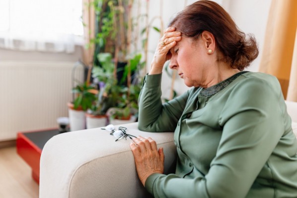 Eine etwa 65 jährige Frau sitzt auf einem Sofa. Sie hat die Augen geschlossen und sie hält eine Hand an ihre Stirn