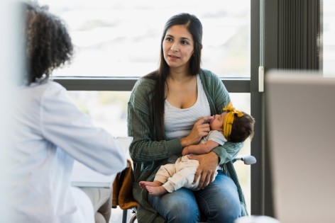 Frau sitzt mit frisch geborenem Baby im Behandlungszimmer ihrer Gynäkologin.