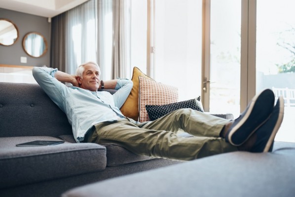 Ein etwa 65-jähriger Mann sitzt entspannt zurückgelehnt und mit hochgelegten Beinen auf seinem Sofa.