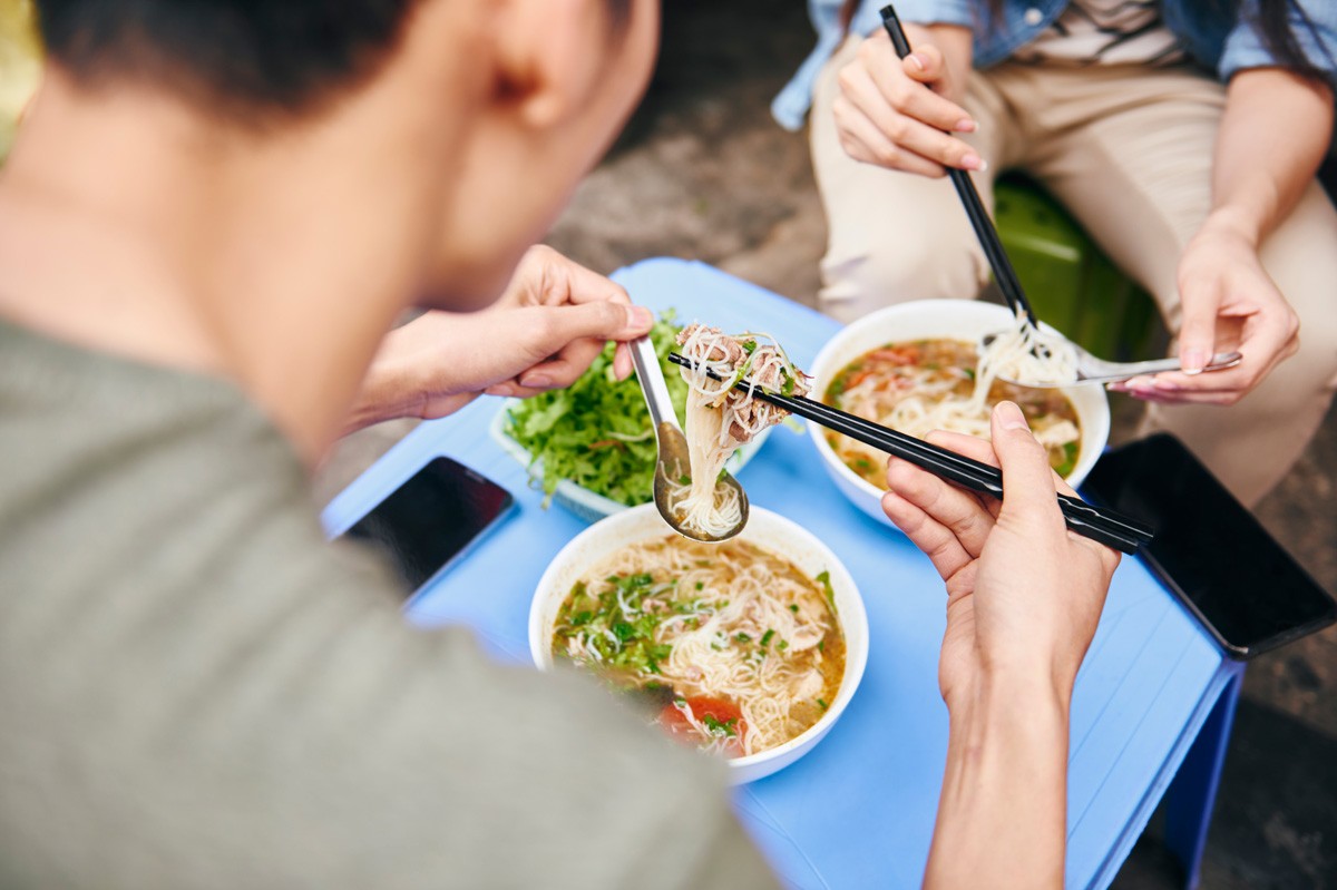 Zwei Männer essen draußen asiatische Suppe. Sie benutzen Stäbchen und Löffel.