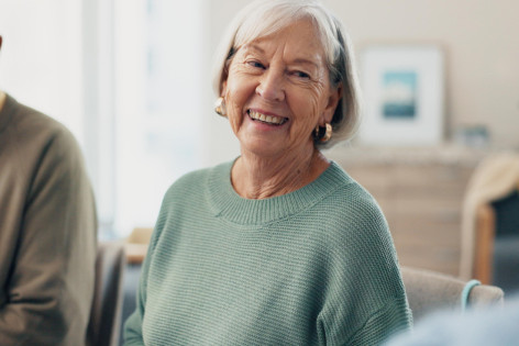 Zwei Seniorinnen sitzen auf einer Couch im Wohnzimmer und trinken gemeinsam Kaffee. Beide sind zueinander gedreht und lächeln sich an.