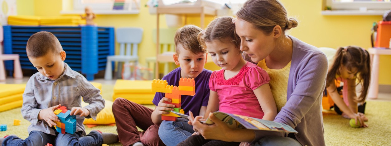 Kita-Erzieherin sitzt auf dem Boden und schaut mit 2 Kindern ein Buch an. Ein weiteres Kind sitzt daneben und spielt mit Bausteinen. 