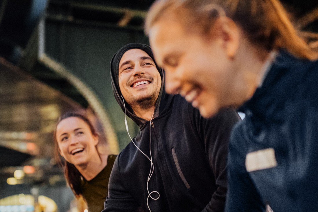 Ein junger Mann und zwei junge Frauen, alle Anfang 20, bereiten sich zusammen aufs gemeinsame Laufen vor. Sie befinden sich zum Abend in urbanem Raum und sind guter Laune.