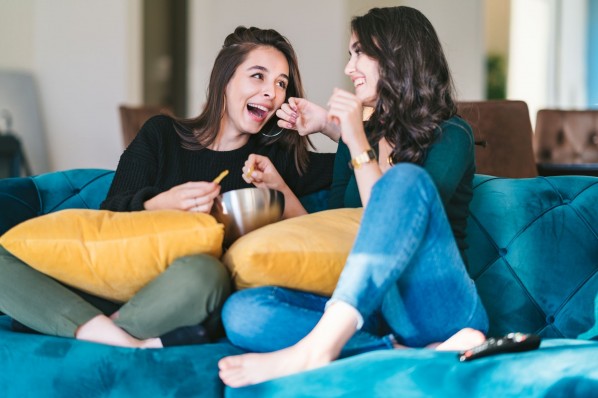 Zwei junge Frauen Anfang 20 sitzen auf dem Sofa, lachen und essen Gemüsechips