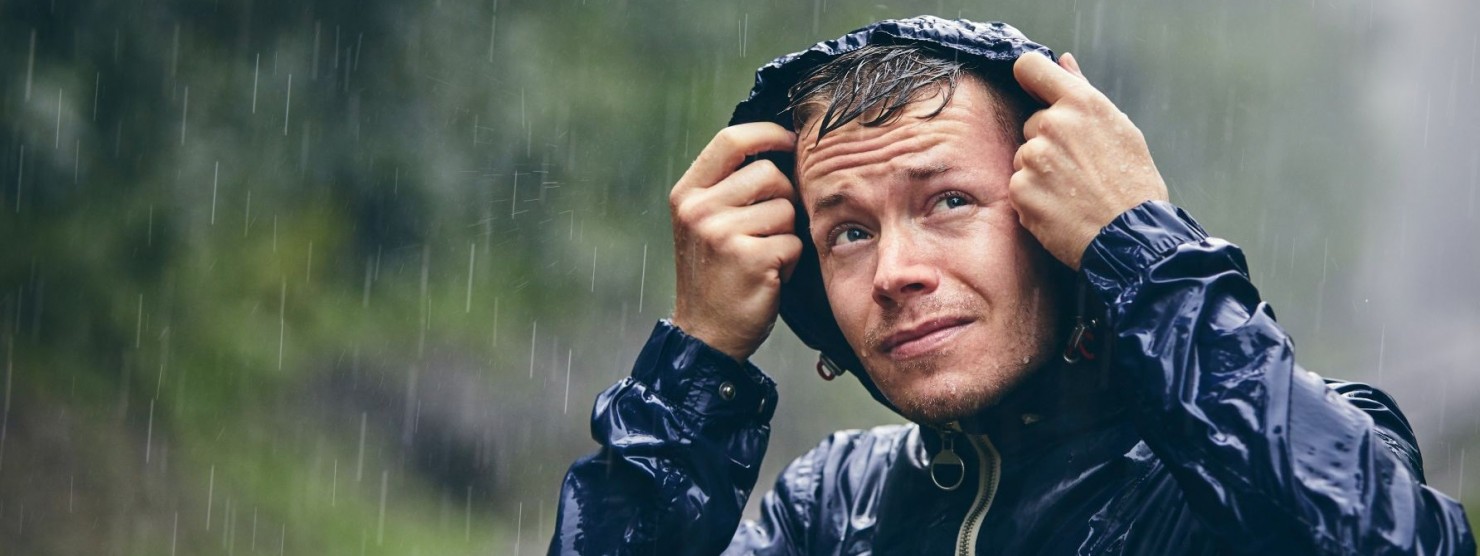Ein etwa 30 jähriger Mann steht in der Natur im strömenden Regen. Er trägt eine Regenjacke und hat die Kapuze über den Kopf gezogen.