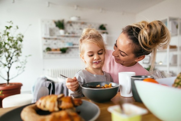 Ein etwa 3-jähriges Mädchen isst Müsli. Sie sitzt auf dem Schoß ihrer Mutter.