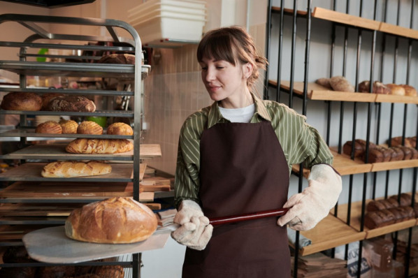 Eine junge Bäckereifachverkäuferin holt frisch gebackene Brotlaibe aus dem Ofen