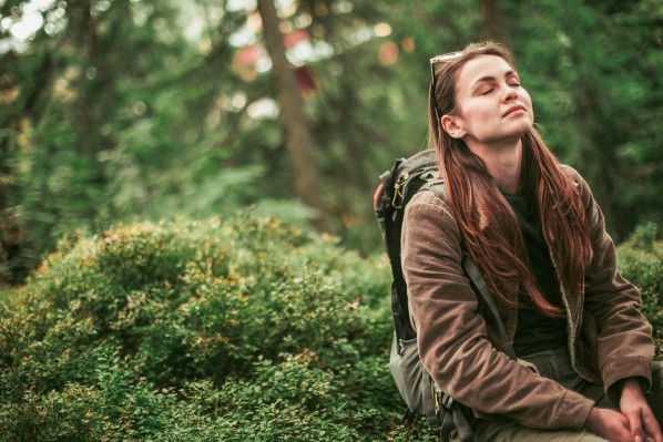 Eine etwa 30-jährige Frau sitzt mit ihrem Wanderrucksack auf einer moosigen Stelle im Wald. Sie lehnt den Kopf zurück, schließt die Augen und lächelt.