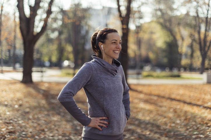 Eine Frau steht lächelnd in einem winterlichen Park. Sie hat Kopfhörer im Ohr und macht sich bereit für Sport.