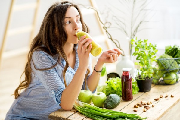 Eine etwa 35 jährige Frau sitzt an einem Tisch und trinkt einen Saft aus einer Plastikflasche. Auf dem Tisch liegen noch weitere gesunde Nahrungsmittel wie Äpfel und Avocado.