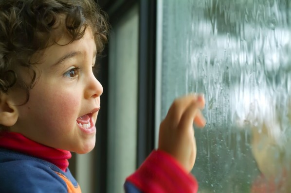 Ein etwa 2 jähriger Junge steht an einem Fenster und fasst an die Glasscheibe. Draußen regnet es.