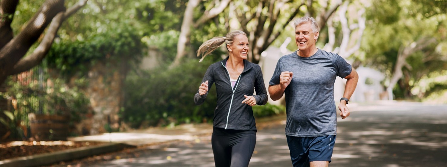 Ein Mann und eine Frau, beide etwa 50, joggen gemeinsam auf einer von Bäumen umsäumten Straße.