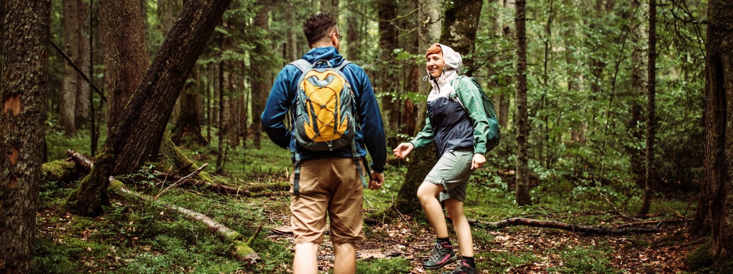 Eine Frau und ein Mann, beide Mitte 20, wandern im Wald. Die Frau dreht sich zu dem Mann um.