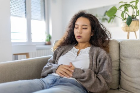 Eine junge Frau sitzt auf ihrem Sofa und leidet an Unterleibsschmerzen.