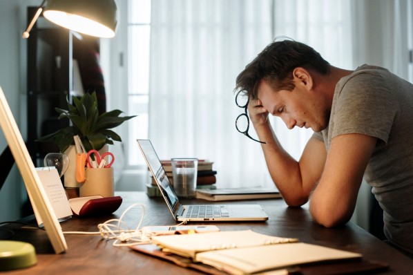 Ein etwa 30 jähriger Mann sitzt am Schreibtisch vor seinem Laptop und stützt seinen Kopf in die rechte Hand. Er sieht gestresst aus.