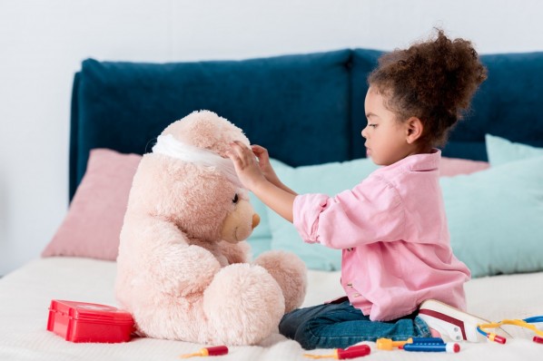 Ein etwa 4 jähriges Mädchen verbindet mit einer Mullbinde den Kopf eines großen Teddybären.
