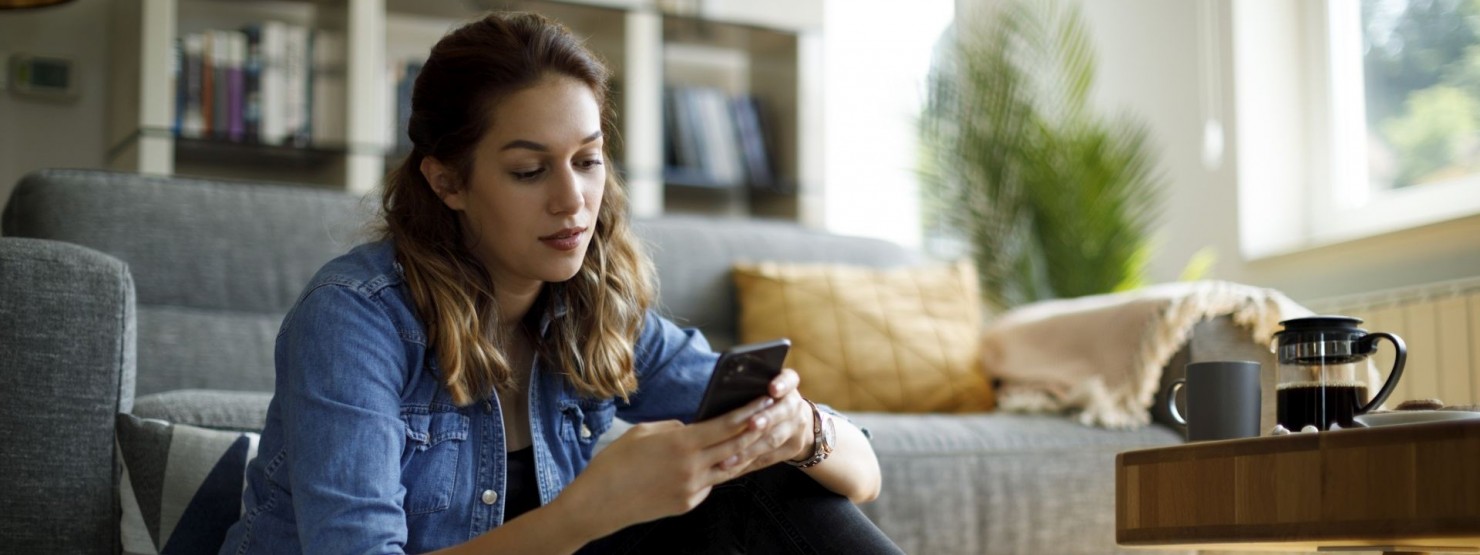 Junge Frau sitzt in ihrem Wohnzimmer auf dem Fußboden und schaut nachdenklich auf ihr Handy. 