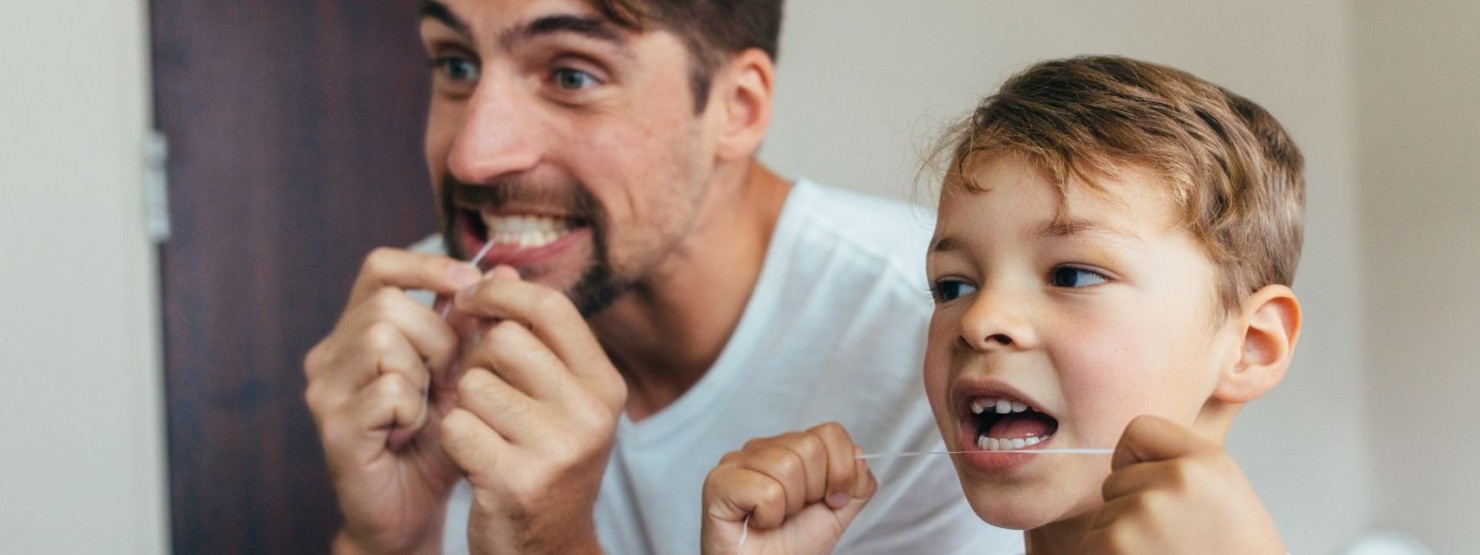 Ein etwa 6-jähriger Junge steht mit seinem Vater im Badezimmer. Sie pflegen mit Zahnseide ihre Zähne.