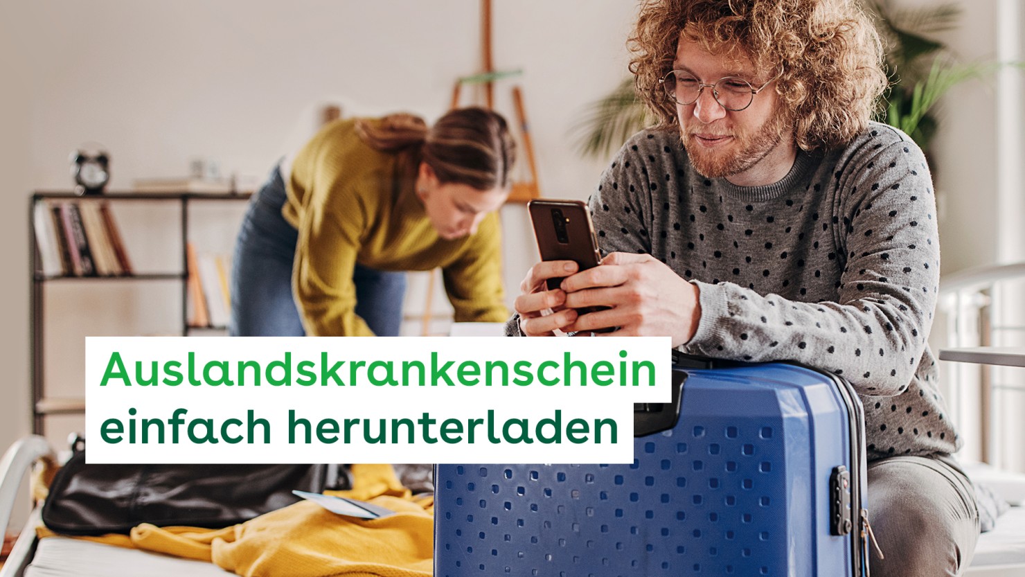Ein Paar, beide etwa 30 Jahre, bereiten eine Reise vor. Er lehnt mit den Armen auf seinem fertig gepackten Koffer und hält ein Smartphone in den Händen. Sie packt im Hintergund ihren Koffer.  