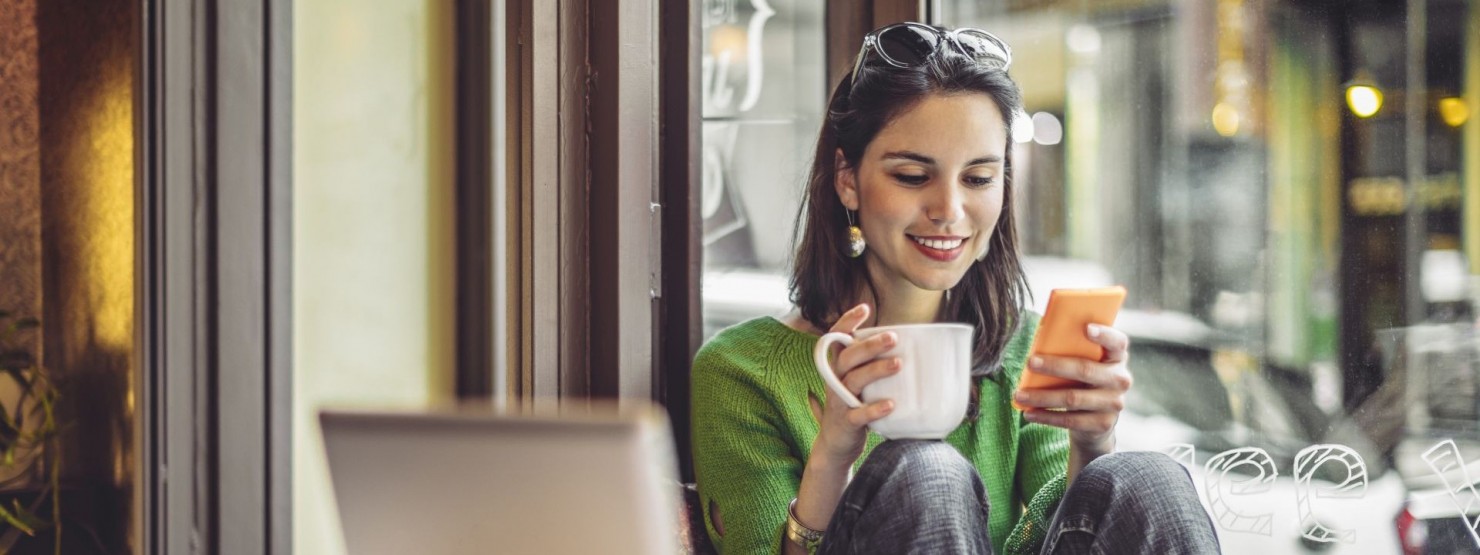 Eine junge Frau sitzt in einem Cafe. Sie schaut lächelnd auf ihr Handy und hält in der rechten Hand eine Tasse. Am Tisch vor ihr steht ein Laptop und Unterlagen.