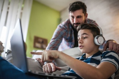 Eine etwa 10 jährige Junge sitzt auf dem Sofa, trägt Kopfhörer und hat den Laptop auf seinem Schoß. Sein Vater steht hinter ihm und schaut ebenfalls auf den Bildschirm. Beide zeigen auf den Laptop.
