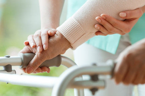 Eine ältere Person nutzt zum laufen eine Gehhilfe und wird von einer zweiten Person gestützt.
