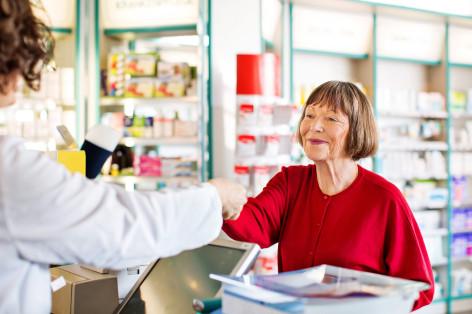 Eine Seniorin in roten Pullover steht in einer Apotheke und lässt sich von einer Apothekerin beraten. Sie lächelt.