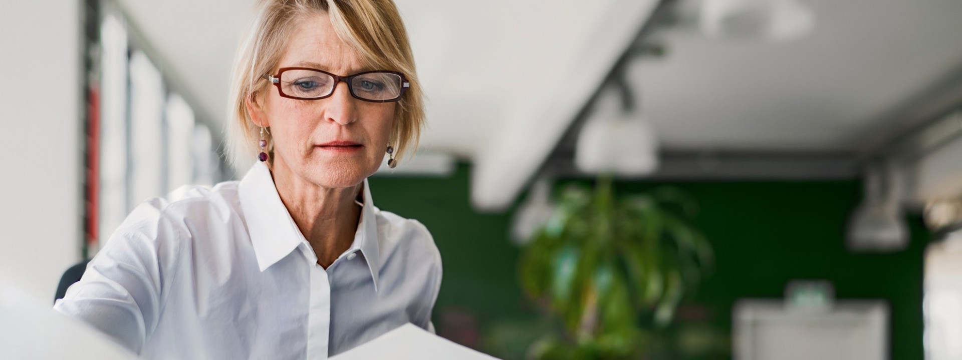 Eine etwa 50-jährige Frau mit blonden Haaren und Brille sortiert an einem Schreibtisch ihre Unterlagen.