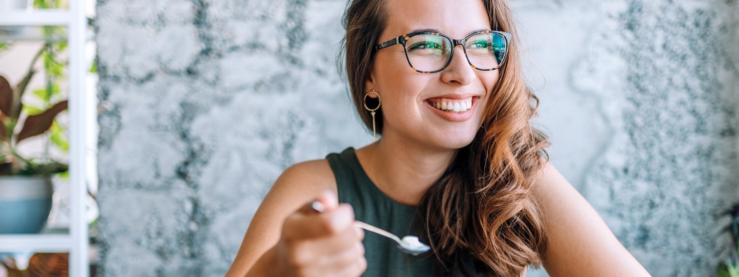 Eine ca. 25 jährige Frau mit Brille und langen Haaren isst einen Naturjoghurt mit Früchten.