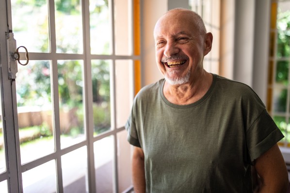 Portrait of casually clothed happy senior man standing near window at home and smilng