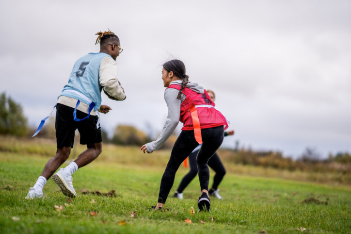 A group of University students run around on a field as they play a game of flag football together. They are each dressed comfortably and have numbered team pinnies on.