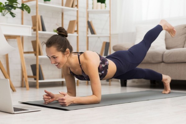 Eine etwa 20-Jährige macht einen Plank mit hoch gestreckten Bein auf Ihrer Sportmatte im Wohnzimmer.